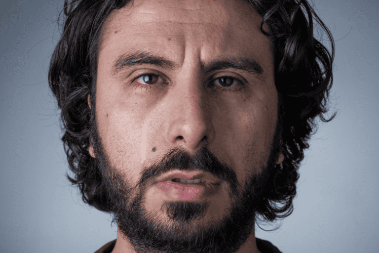 A contemplative man with wavy dark hair and a full beard, gazing intensely with a serious expression against a neutral gray background.
