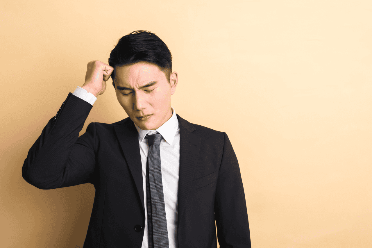Feeling confused or overwhelmed, young businessman in formal suit holding head with hand, standing against beige background.