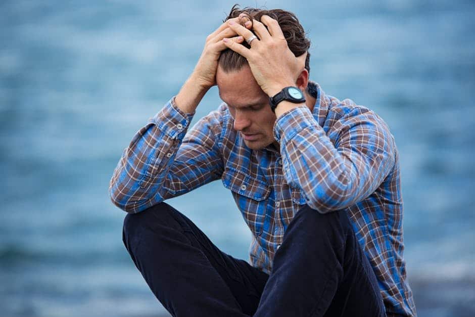 Caught in emotional distress, man sitting by the water with hands on his head, experiencing stress or sadness, casual clothing, outdoor setting, emphasizing mental health and emotional struggles.