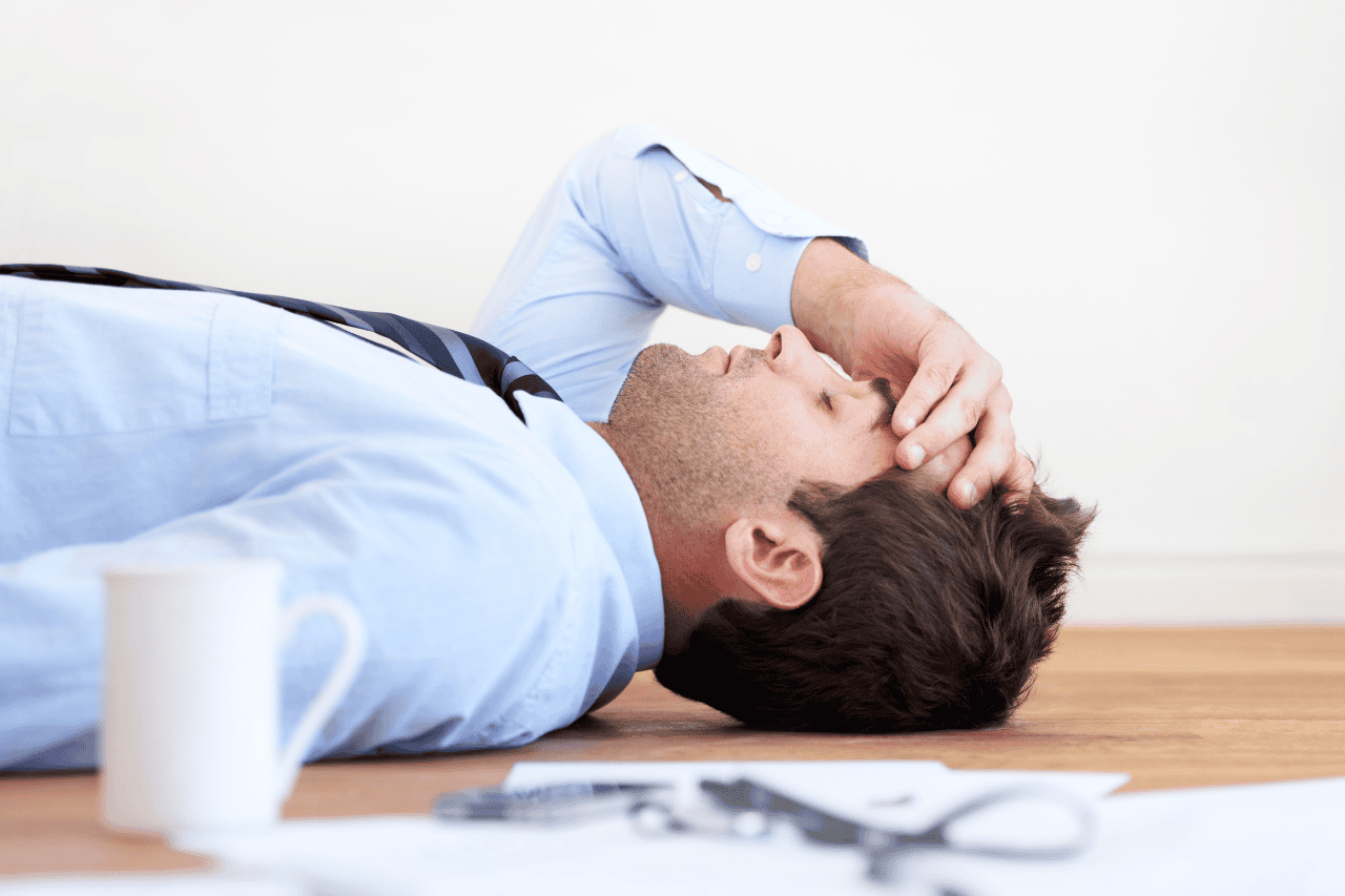 Stressed businessman in formal attire lying on the floor with his hand on his forehead, experiencing fatigue or anxiety, in a professional office setting.