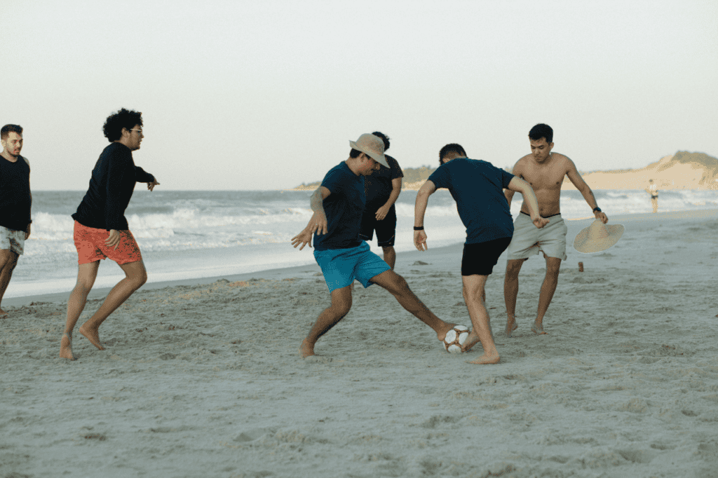 Beach soccer game with young men playing barefoot near shoreline on sandy beach during daytime.