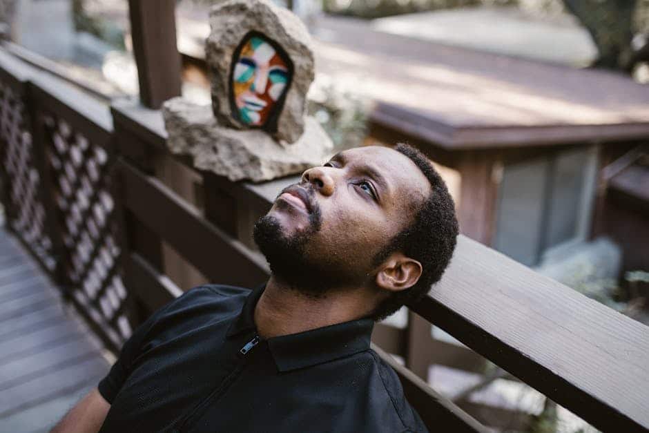 Vegan man relaxing outdoors on a wooden deck with a colorful abstract face sculpture in the background, emphasizing his contemplative mood and modern art decor.