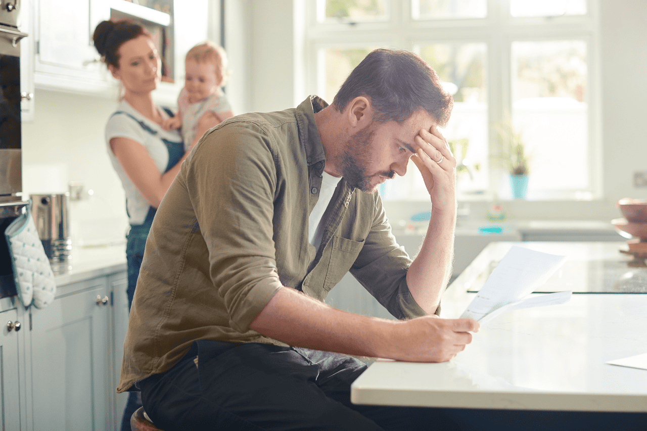 Frustrated man reading a letter at kitchen counter with a woman and child in the background, portraying emotional stress and family dynamics - The Solemn Sir.