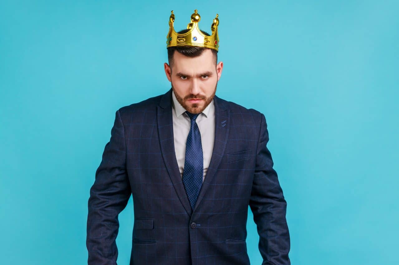 Confident man wearing a navy business suit with a crown on his head, symbolizing leadership and authority, against a bright blue background, representing the regal and powerful theme of The Solemn Sir.