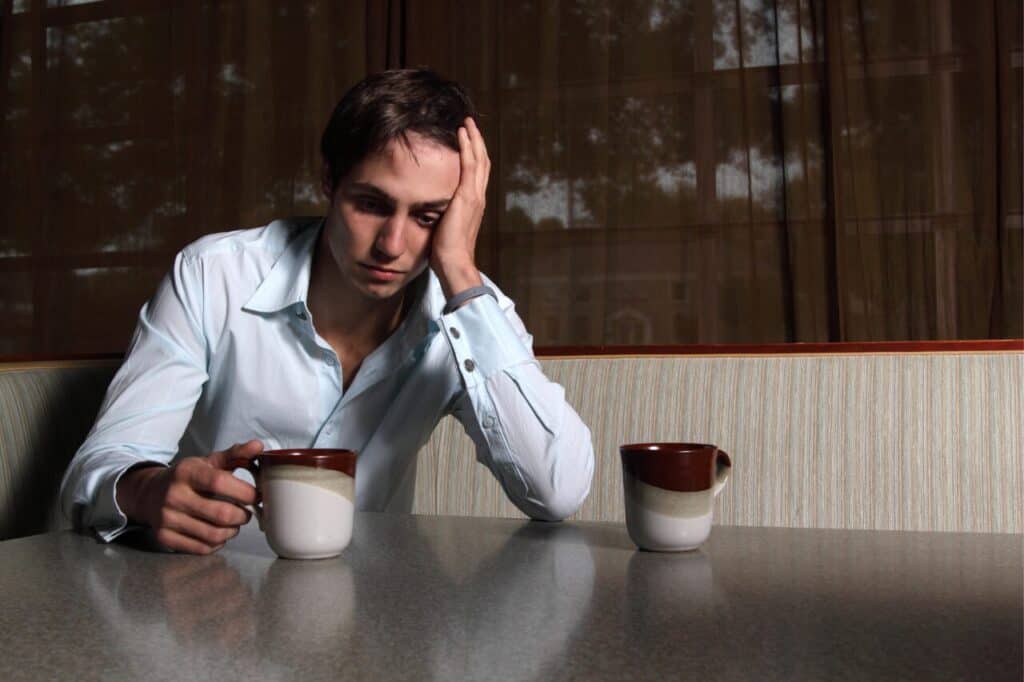A stressed young man sitting at a table with two coffee mugs, holding one mug while resting his head on his hand, expressing fatigue or frustration.