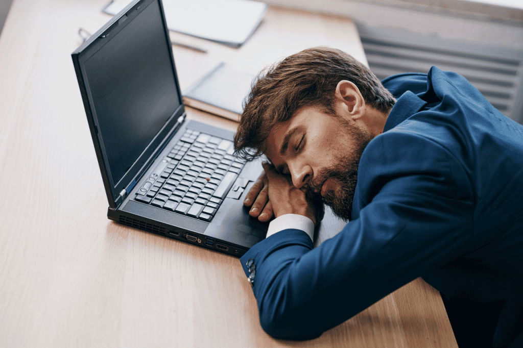 Tired businessman sleeping at desk with laptop, feeling exhausted after work, representing burnout or need for rest and self-care.