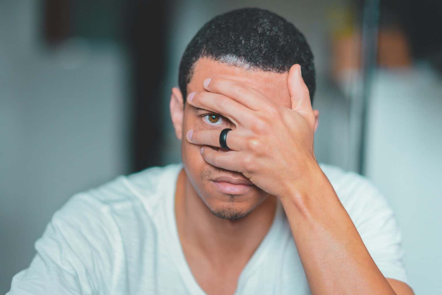 A close-up portrait of an adult man with one eye covered by his hand, wearing a black ring.