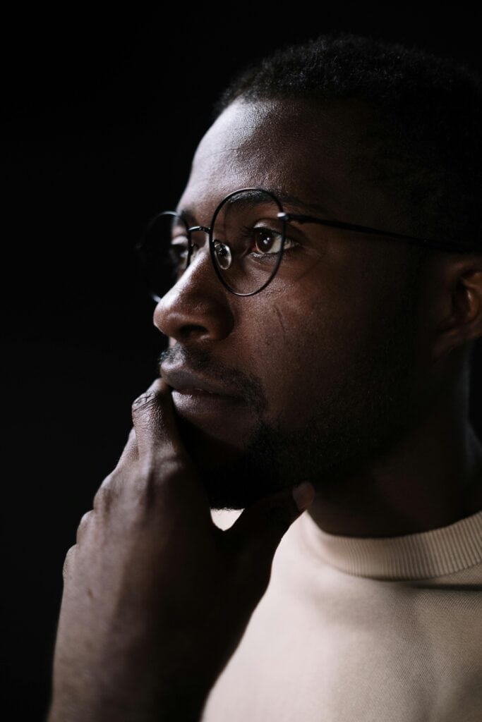 Close-up portrait of a thoughtful man wearing round glasses against a dark background.