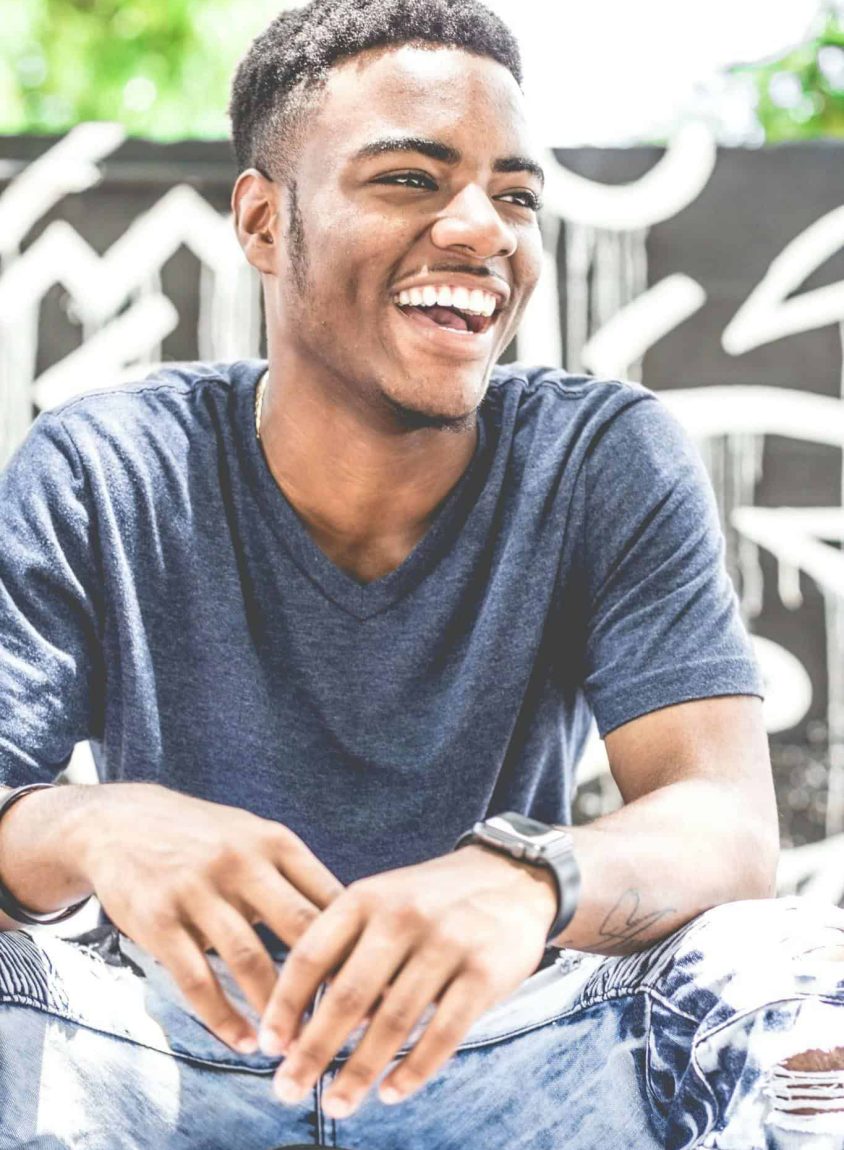 Happy young man sitting outdoors with a bright smile, wearing casual clothing. Ideal for lifestyle and fashion themes.